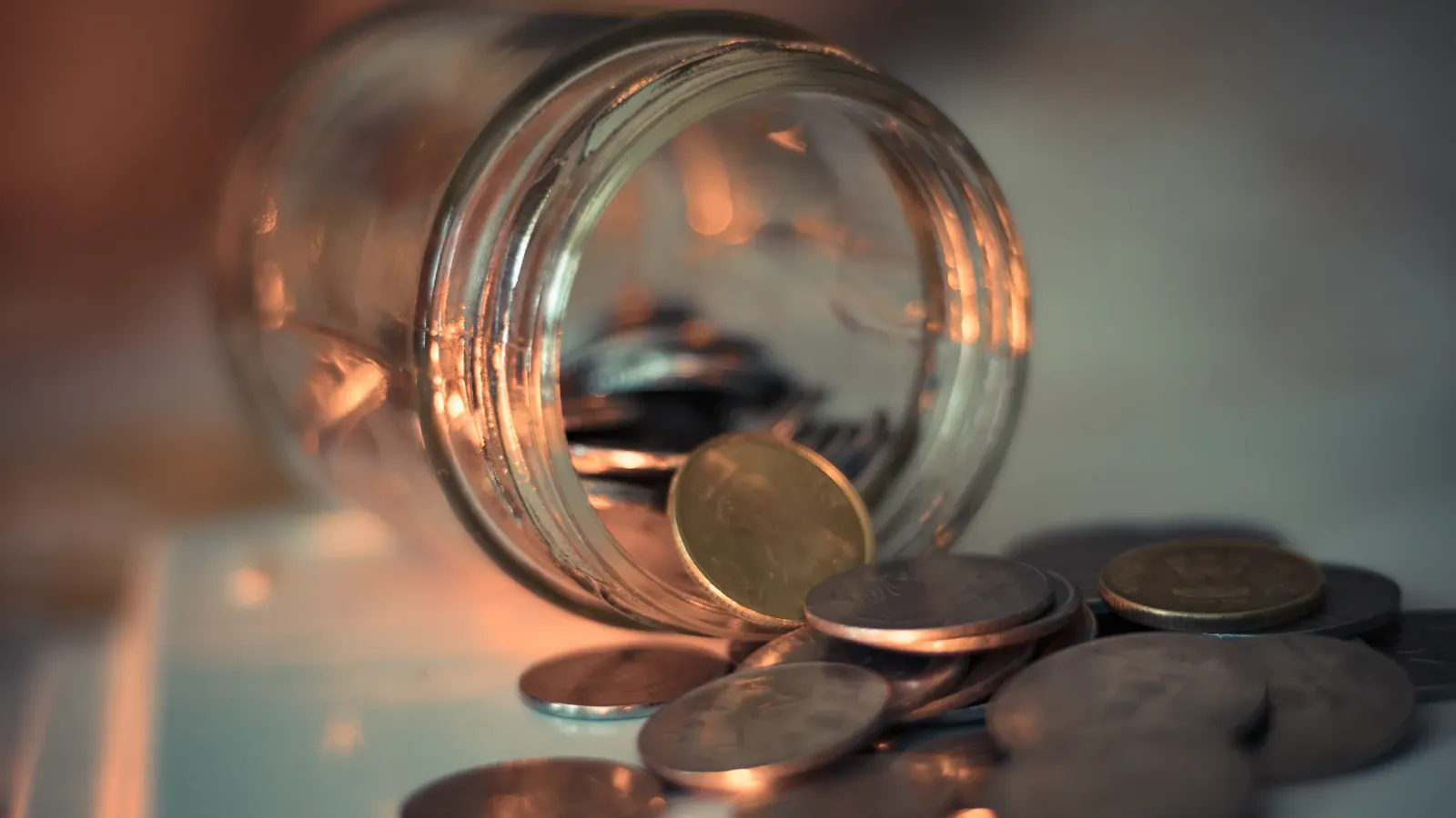 A tipped-over glass jar with coins spilling onto a table