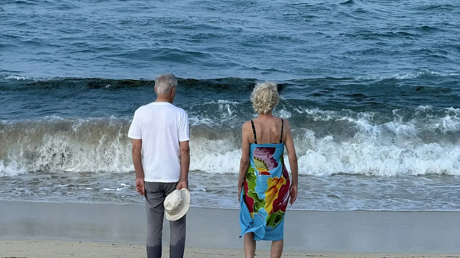 An older couple standing on a beach, looking out at the ocean waves