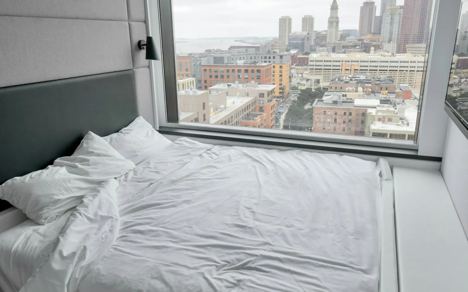 A hotel bed beside a large window with a city view